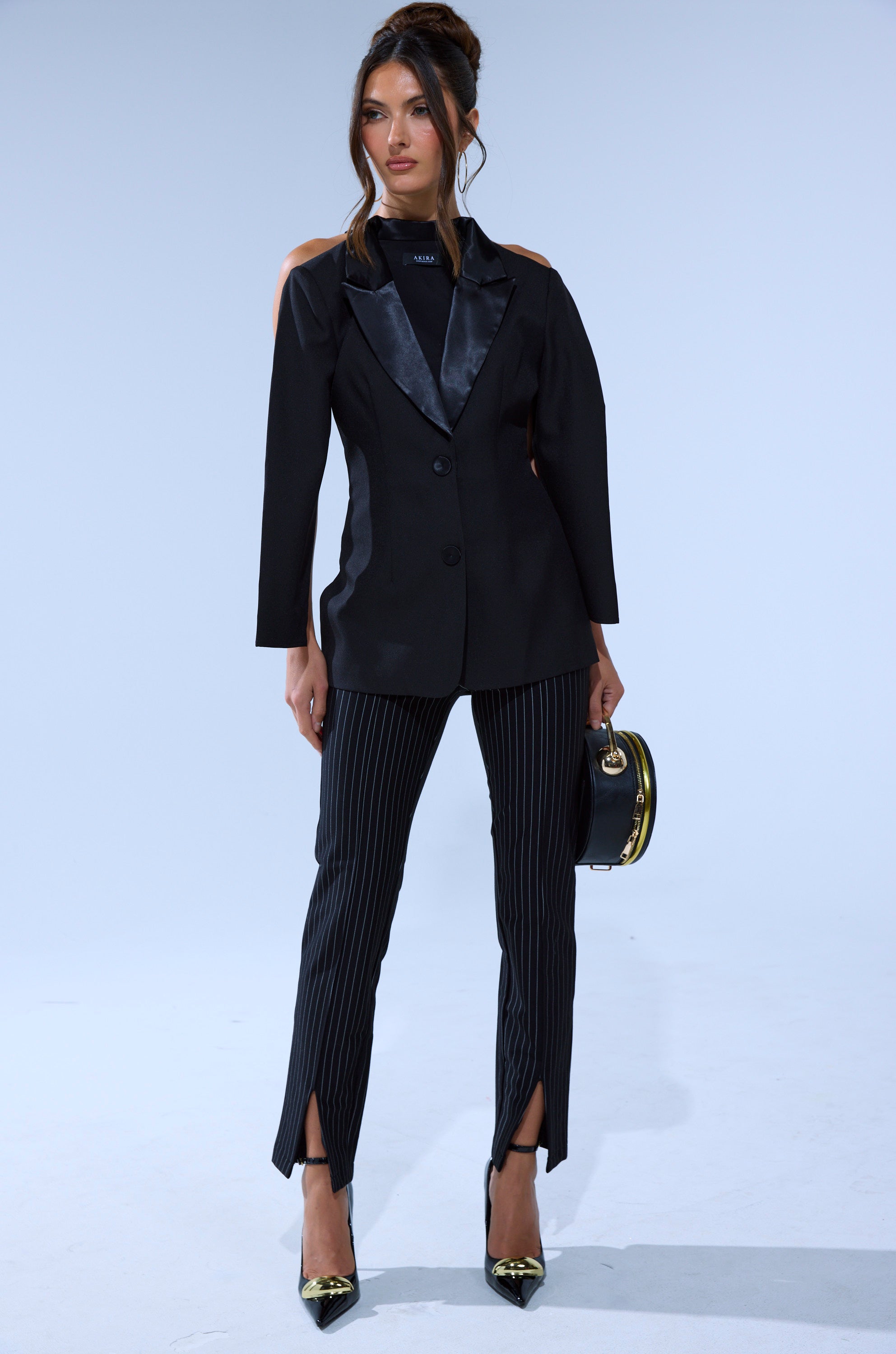 A woman stands against a light background wearing LIVI Stretchy Pinstripe Skinny Pants with slits, a black blazer with satin lapels, pointed black heels with gold tips, and holds a small black handbag. Her hair is styled in an updo.