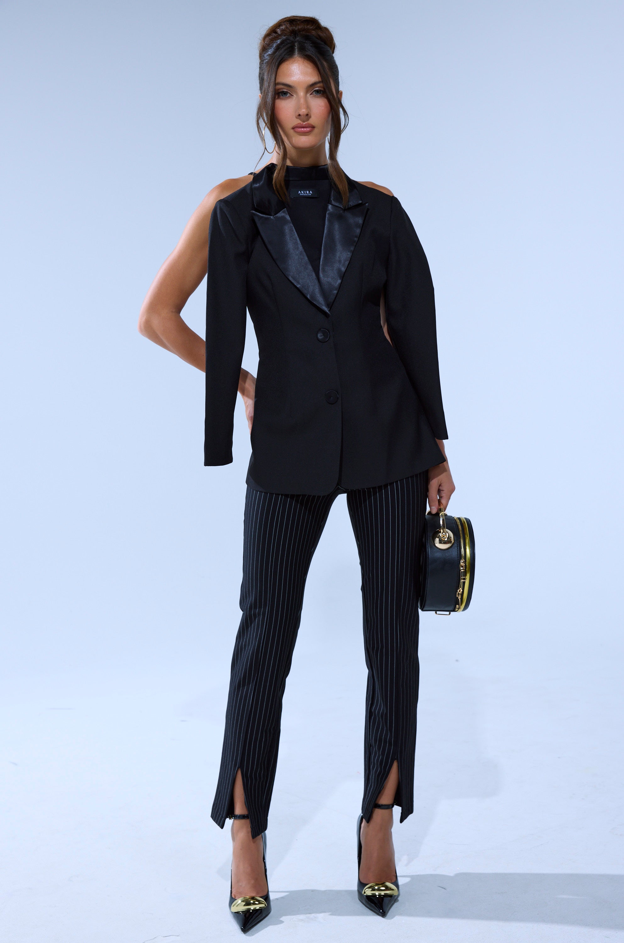 A woman poses against a plain background wearing black satin lapel blazer, LIVI STRETCHY PINSTRIPE SKINNY PANTS WITH SLITS, black high heels with gold tips, and carrying a round black handbag.