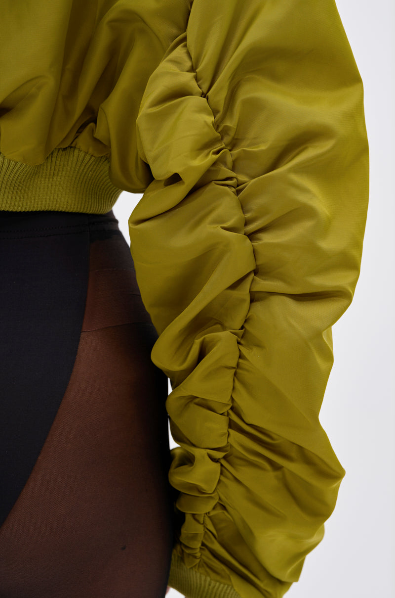 A close-up of a person wearing the TAKE IT EASY CROPPED BOMBER IN GREEN, highlighting its ruched sleeve detail and cropped fit with black sheer tights—perfect for transitional layering.