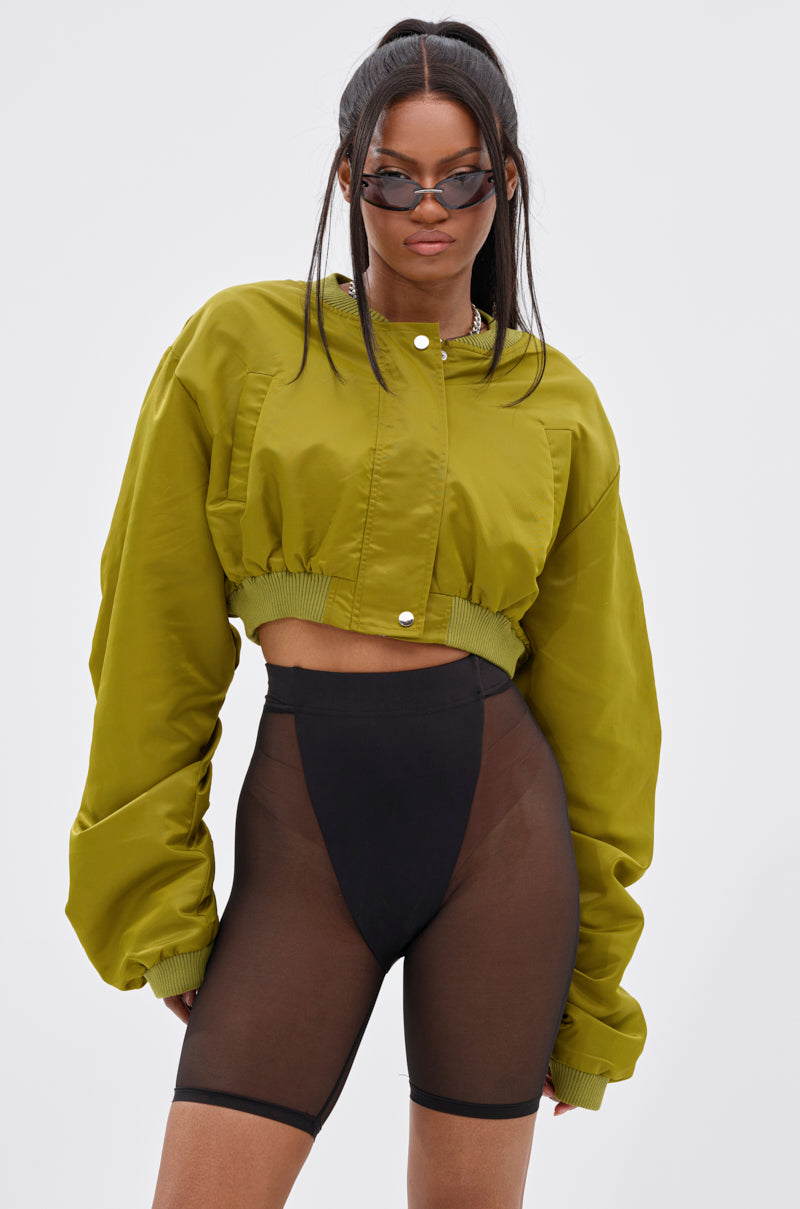 A woman models the TAKE IT EASY CROPPED BOMBER IN GREEN with black sheer high-waisted shorts, black undergarments, and sunglasses against a light gray background—a stylish outfit for transitional layering.