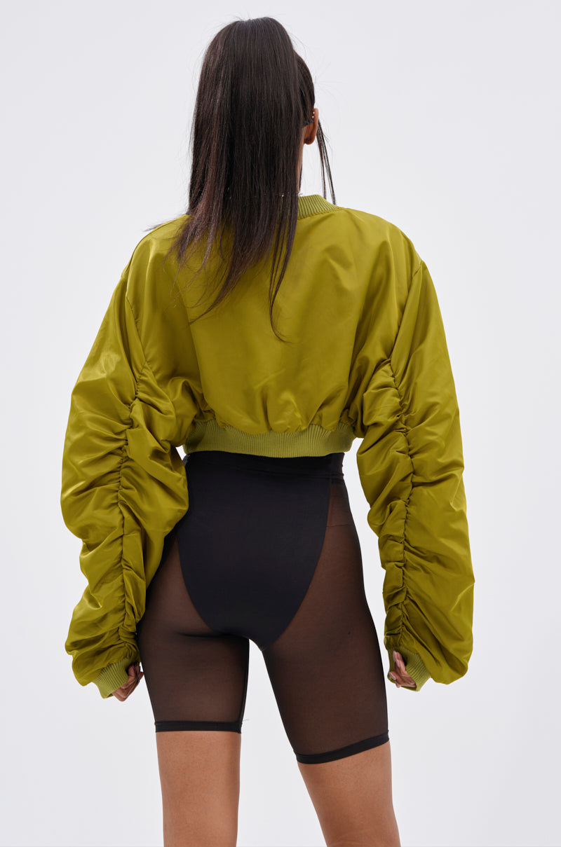 Wearing the TAKE IT EASY CROPPED BOMBER IN GREEN, a person with long dark hair and ruched sleeves stands with their back to the camera, paired with high-waisted sheer black shorts against a plain light background.