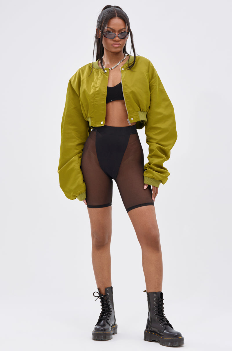 A woman showcases transitional layering in the TAKE IT EASY CROPPED BOMBER IN GREEN, styled with a black bralette, high-waisted sheer black shorts over black underwear, lace-up boots, sunglasses, and a necklace against a plain background.
