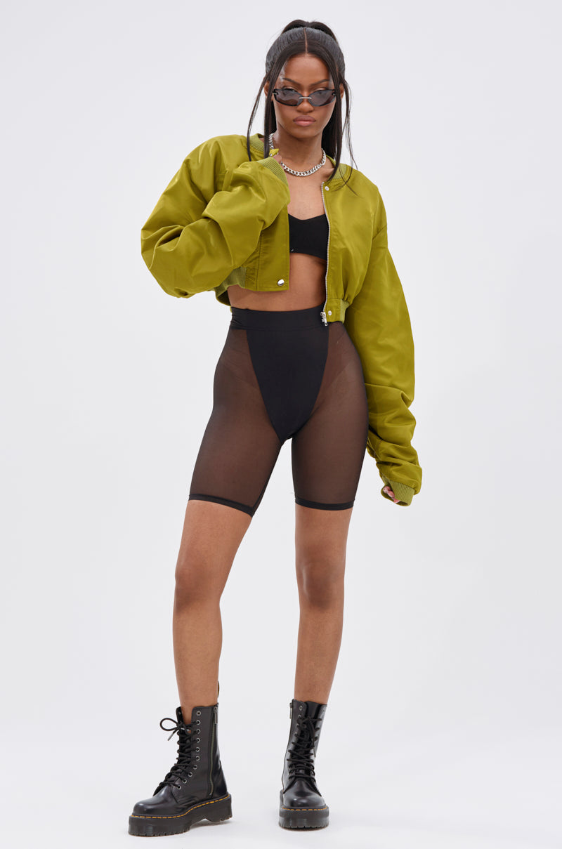 A woman in the TAKE IT EASY CROPPED BOMBER IN GREEN, black sunglasses, chunky necklace, black bra, sheer high-waisted black shorts, and lace-up boots stands confidently against a plain white background.