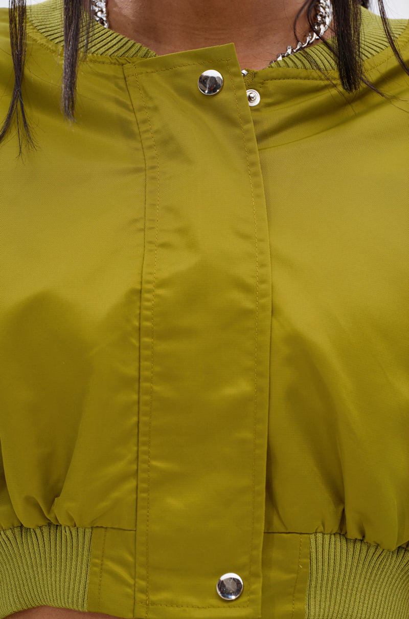 Close-up of a person wearing the TAKE IT EASY CROPPED BOMBER IN GREEN with metal snap buttons and an elastic waistband; a silver chain necklace peeks at the neck—perfect for easy transitional layering.