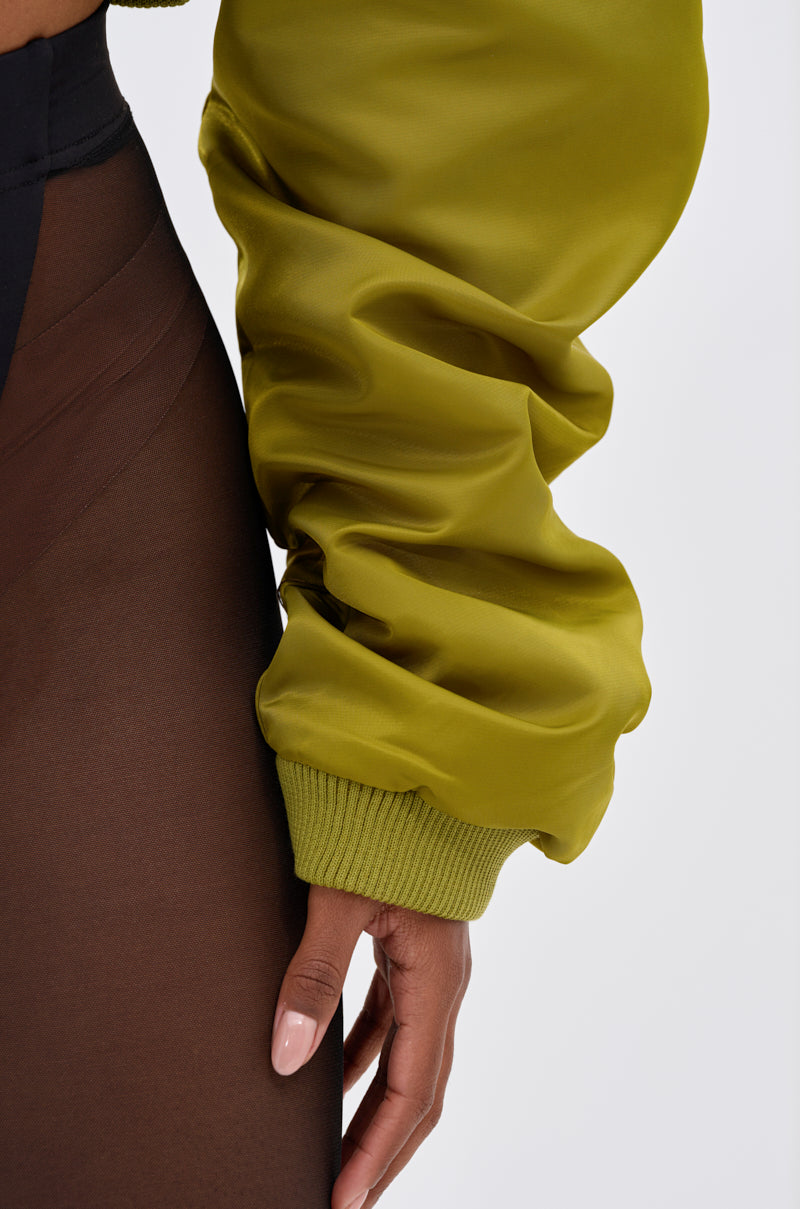 A close-up of a manicured hand wearing the TAKE IT EASY CROPPED BOMBER IN GREEN, styled over a sheer black skirt. The look highlights transitional layering with ribbed cuffs against a plain white background.