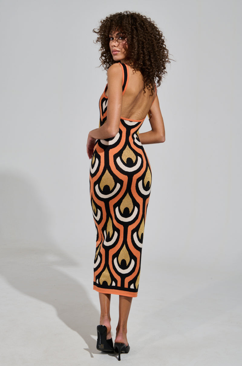 A person with curly hair models the Ziggy Midi Dress in Orange, a sleeveless and backless piece featuring an Art Deco print in orange, black, and beige. Posed sideways in black heels, they highlight the dress's curve-hugging silhouette while gazing into the camera.