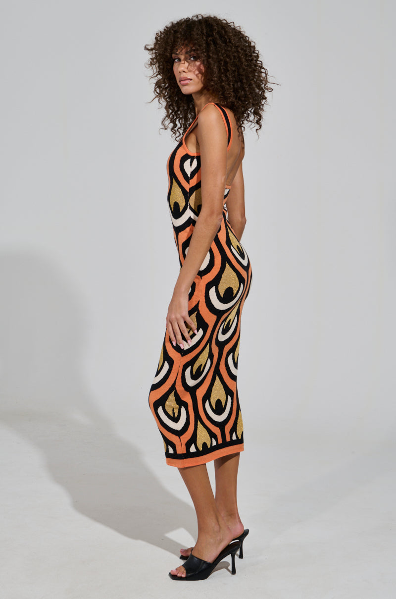 A person with curly hair poses in the ZIGGY MIDI DRESS IN ORANGE, which features a colorful pattern of orange, black, and tan swirls. They showcase the dress's curve-hugging silhouette while standing sideways in black heeled sandals against a simple light background.