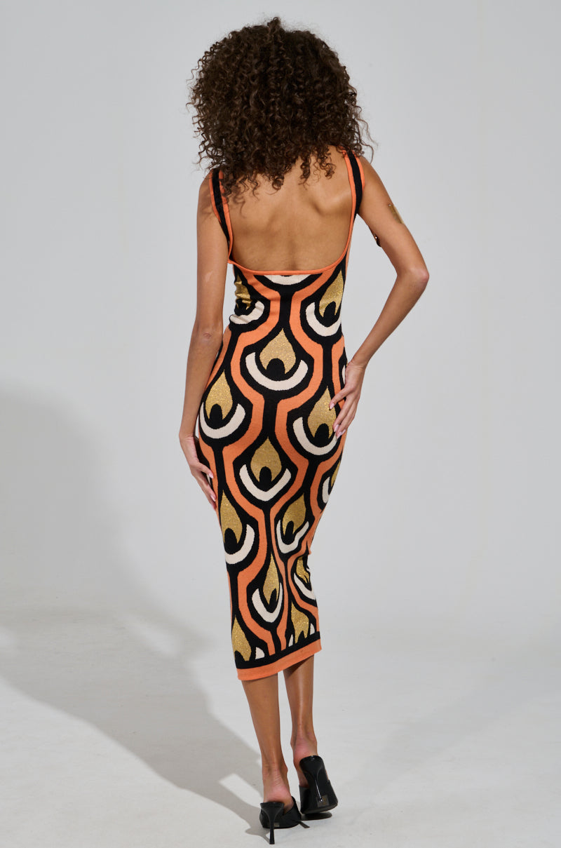 A person with curly hair wears the Ziggy Midi Dress in Orange, a sleeveless and backless piece featuring an Art Deco design in striking black, orange, and yellow. With one hand on their hip and facing away from the camera, they pose to highlight the dress's curve-hugging silhouette while wearing high-heeled shoes against a plain backdrop.