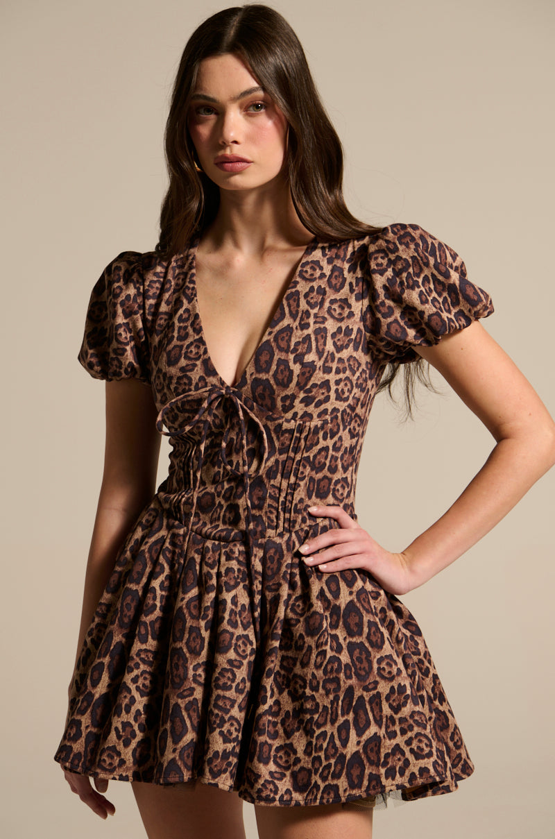 A woman models the WILD THING PRINTED MINI DRESS, highlighting its striking leopard print and stylish puff sleeves. Her long, dark hair enhances the V-neckline as she poses confidently with one hand on her hip against a neutral backdrop.
