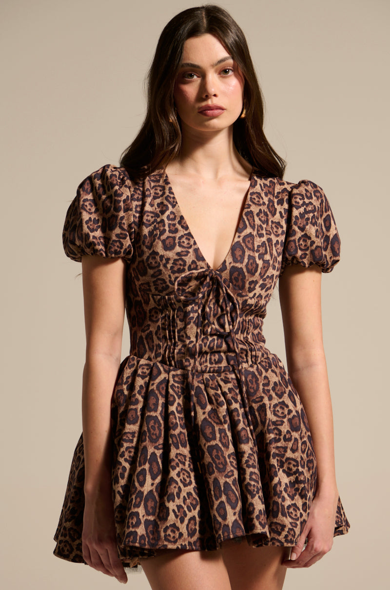 A woman with long hair wears the WILD THING PRINTED MINI DRESS with puff sleeves and a fitted waist against a neutral background. Her leopard-print outfit matches her neutral expression as she faces the camera.