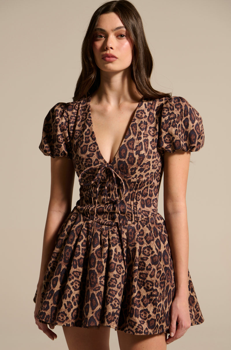 A person with long hair models the WILD THING PRINTED MINI DRESS, highlighting its leopard print and puff sleeves. Featuring a deep V-neckline with laces, they pose confidently against a neutral background.