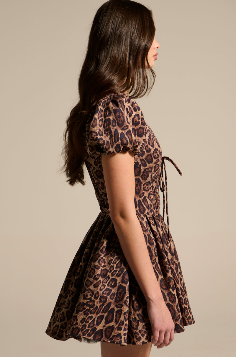 A woman in profile wears the WILD THING PRINTED MINI DRESS, showcasing puff sleeves and a cinched waist. Her long, wavy hair cascades past her shoulders against a plain beige background.