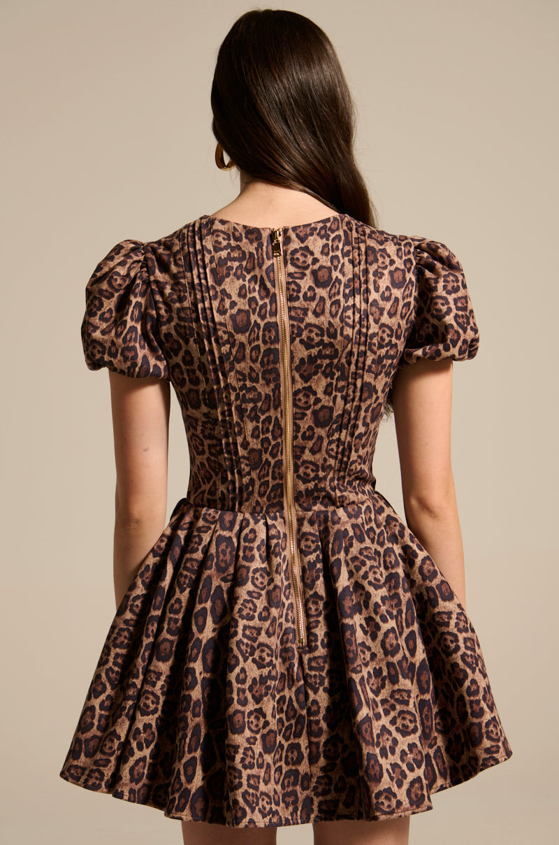 A person with long brown hair, facing away, wears a WILD THING PRINTED MINI DRESS. This leopard print dress features short sleeves, a back zipper, a fitted bodice, and a flared skirt against a plain beige background.