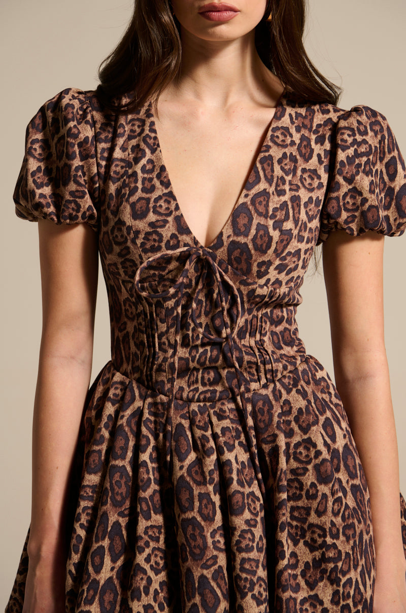 A woman wears the WILD THING PRINTED MINI DRESS, highlighting a leopard print with puff sleeves and a deep V-neckline. It has a fitted bodice, gathered waist, and flared skirt on a neutral beige background.