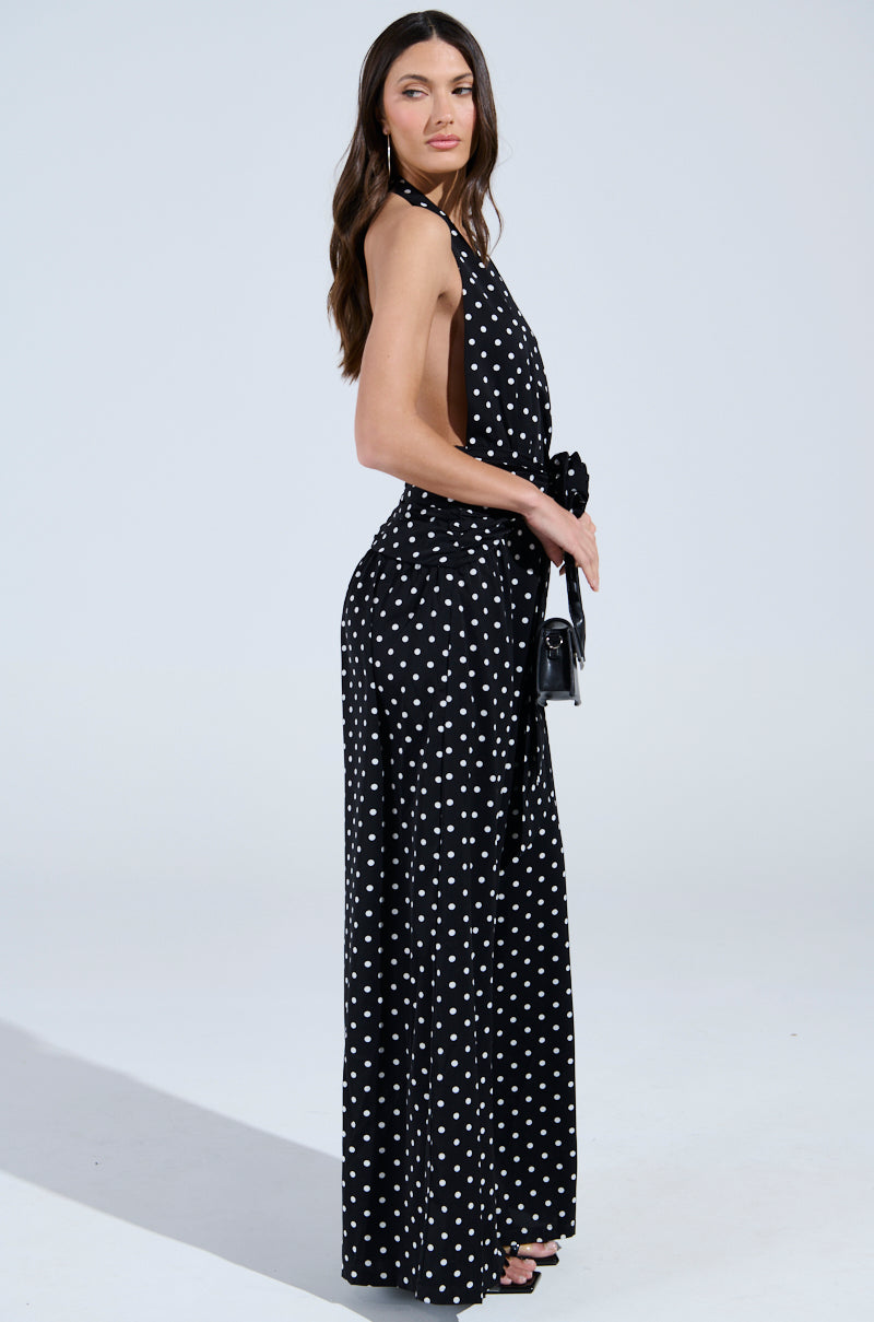 Woman poses sideways against a plain background, wearing the BEST DRESSED POLKA DOT JUMPSUIT—a retro-chic, sleeveless halter-neck style—paired with a small black purse, black high-heeled sandals, and long wavy hair.