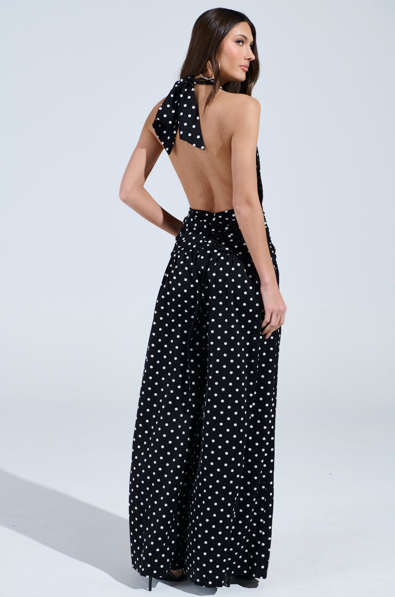 A woman faces away, highlighting the BEST DRESSED POLKA DOT JUMPSUIT with retro-chic polka dots, wide-leg pants, and a sleeveless, backless design; her long brown hair contrasts a simple light background.