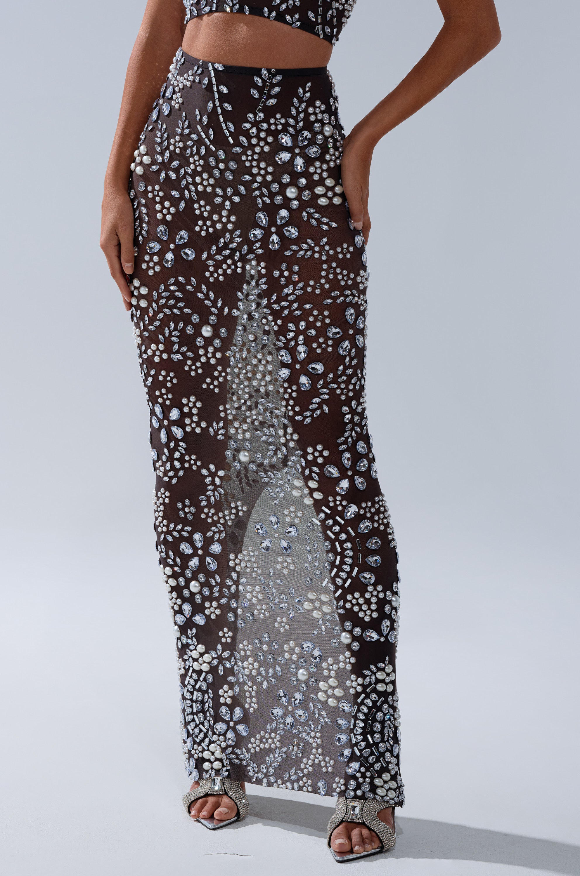 A person wears the BLING BLING MESH RHINESTONE MAXI SKIRT with silver and white pearl embellishments, styled with strappy high-heeled sandals, standing against a plain background.