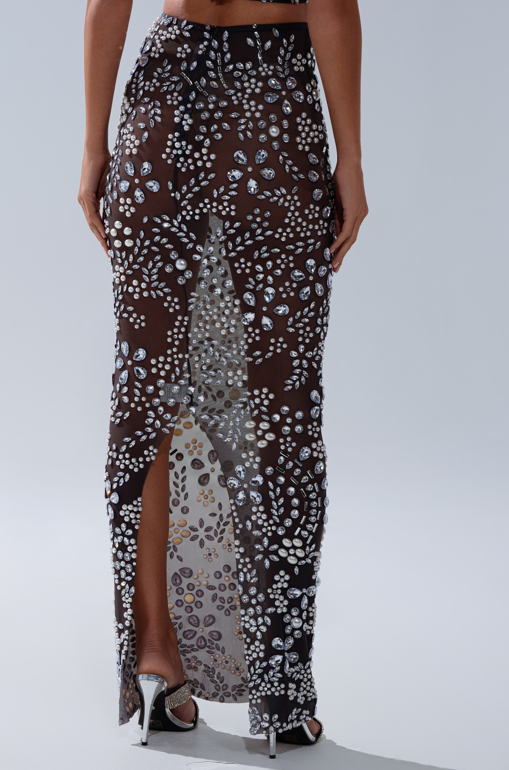 A woman wears the BLING BLING MESH RHINESTONE MAXI SKIRT—a sheer black mesh skirt embellished with sparkling silver rhinestones and floral patterns, featuring a high back slit, paired with high-heeled sandals.