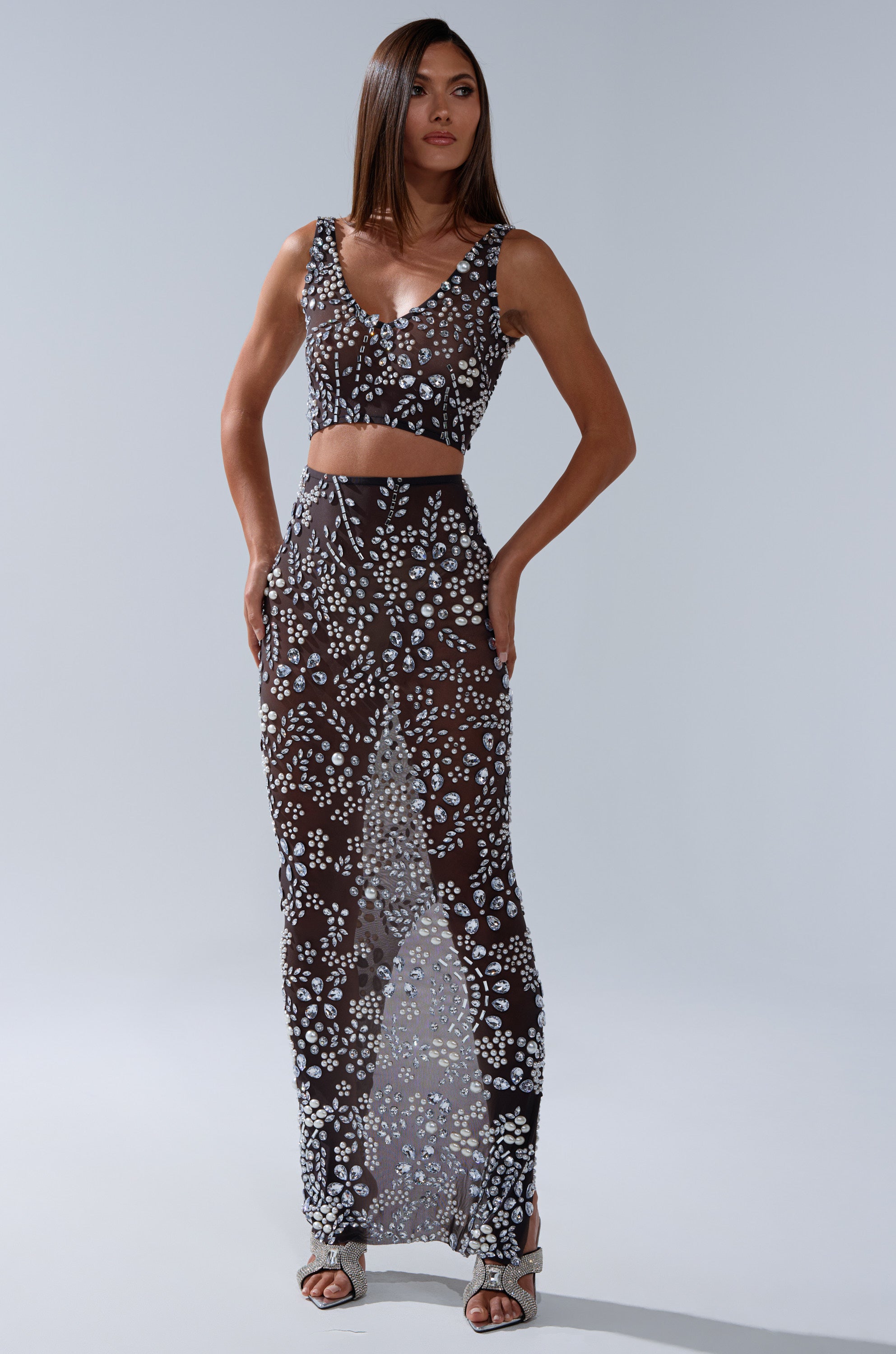 A woman poses in the BLING BLING MESH RHINESTONE BRALETTE, a sleeveless black sheer two-piece with white floral patterns and a cropped fit. She pairs it with silver heeled sandals against a plain light gray background.