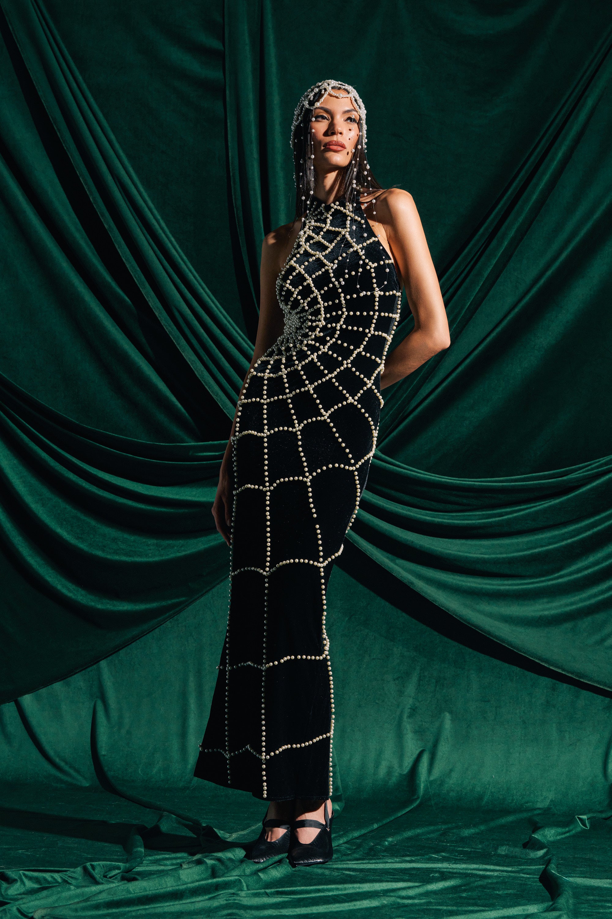 A woman stands confidently before dark green drapes, wearing the MARYJANE MAXI DRESS WITH PEARLS and a matching beaded headpiece, striking a pose with one hand on her hip.