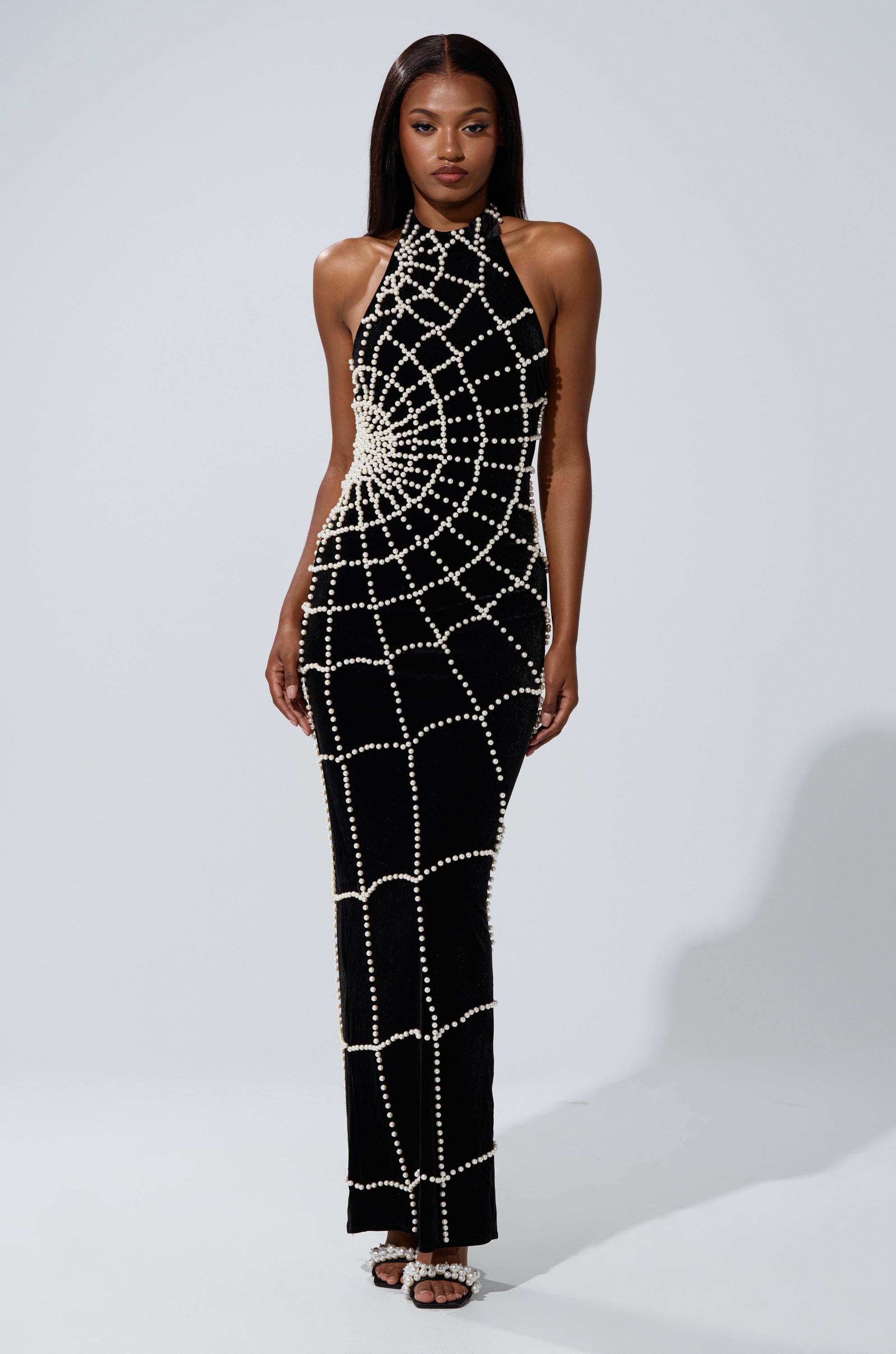 A woman wears the sleeveless black MARYJANE MAXI DRESS WITH PEARLS, featuring spider web pearl embellishments, standing against a plain light gray background.