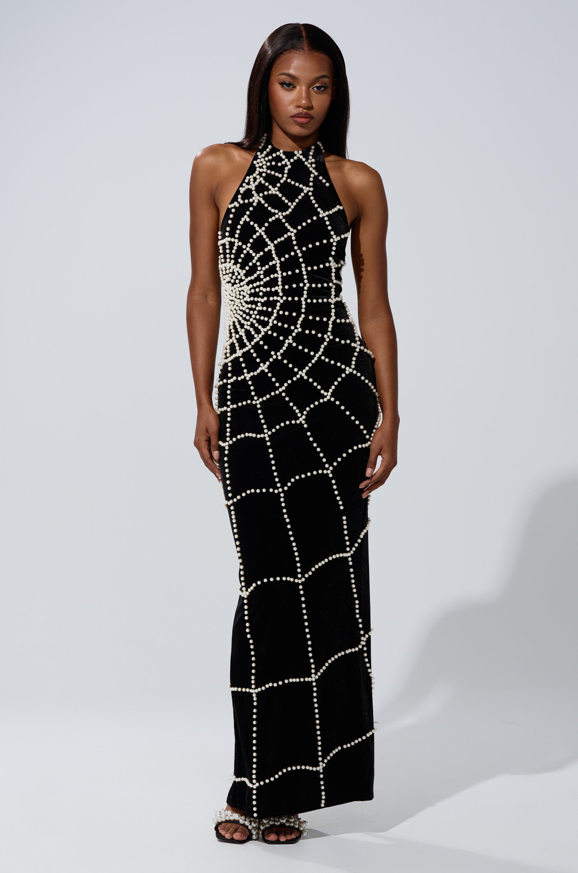 A woman wears the MARYJANE MAXI DRESS WITH PEARLS—a long black halter-neck gown featuring a web-like pattern of white pearls. She poses with straight hair and neutral makeup against a plain light background.