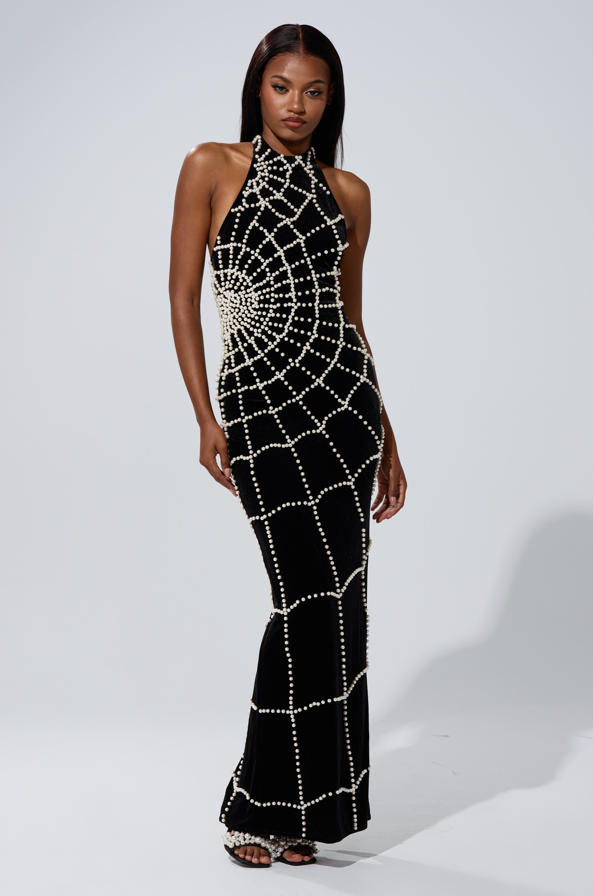 A woman stands confidently in the MARYJANE MAXI DRESS WITH PEARLS, a sleeveless, floor-length black gown adorned with pearl-beaded spiderwebs. Her straight dark hair and sandals complete the look against a plain light gray background.