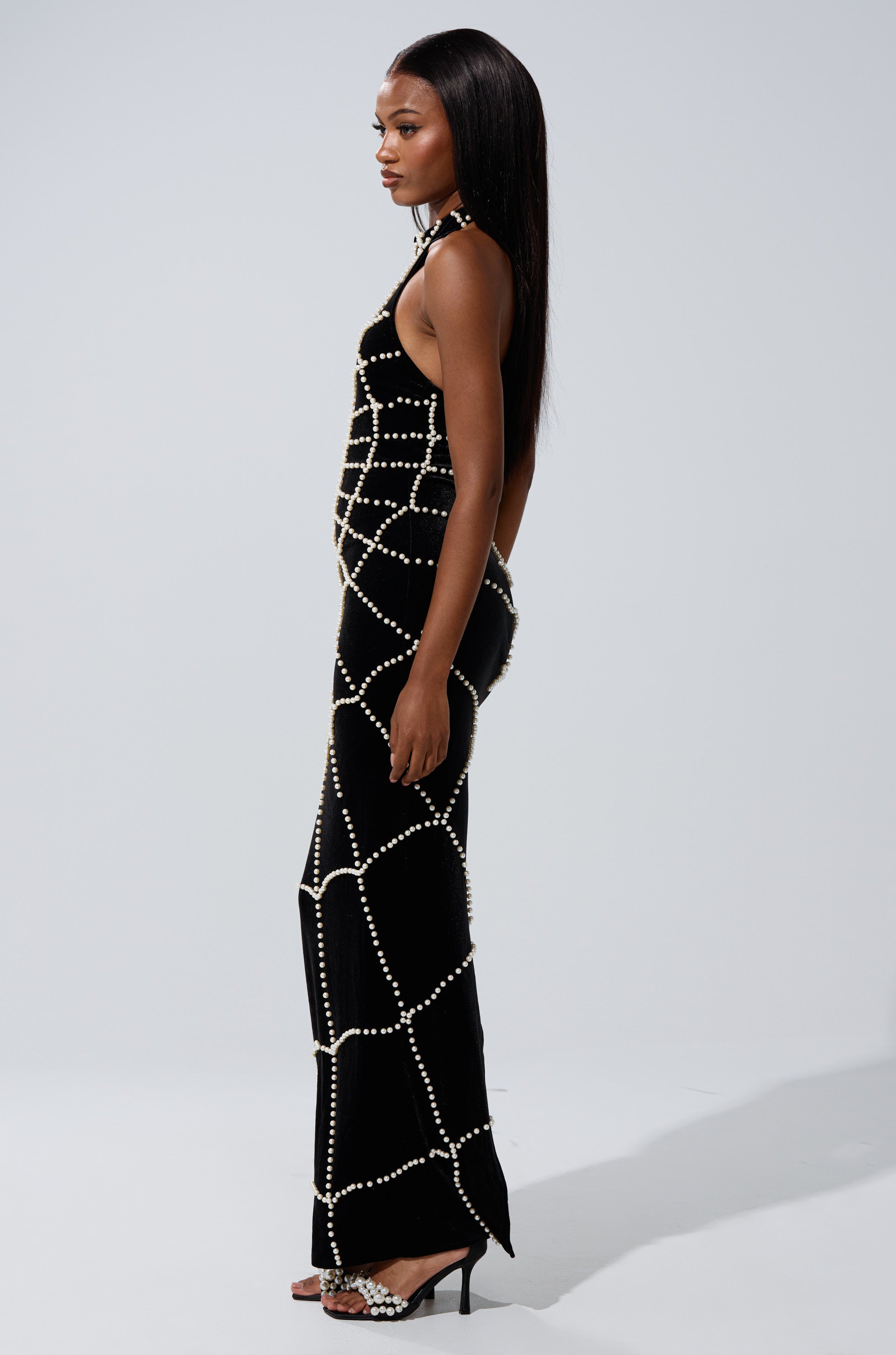 A woman in profile wears the form-fitting black MARYJANE MAXI DRESS WITH PEARLS, styled with high-heeled sandals. Her long, straight hair and neutral expression complete the look.