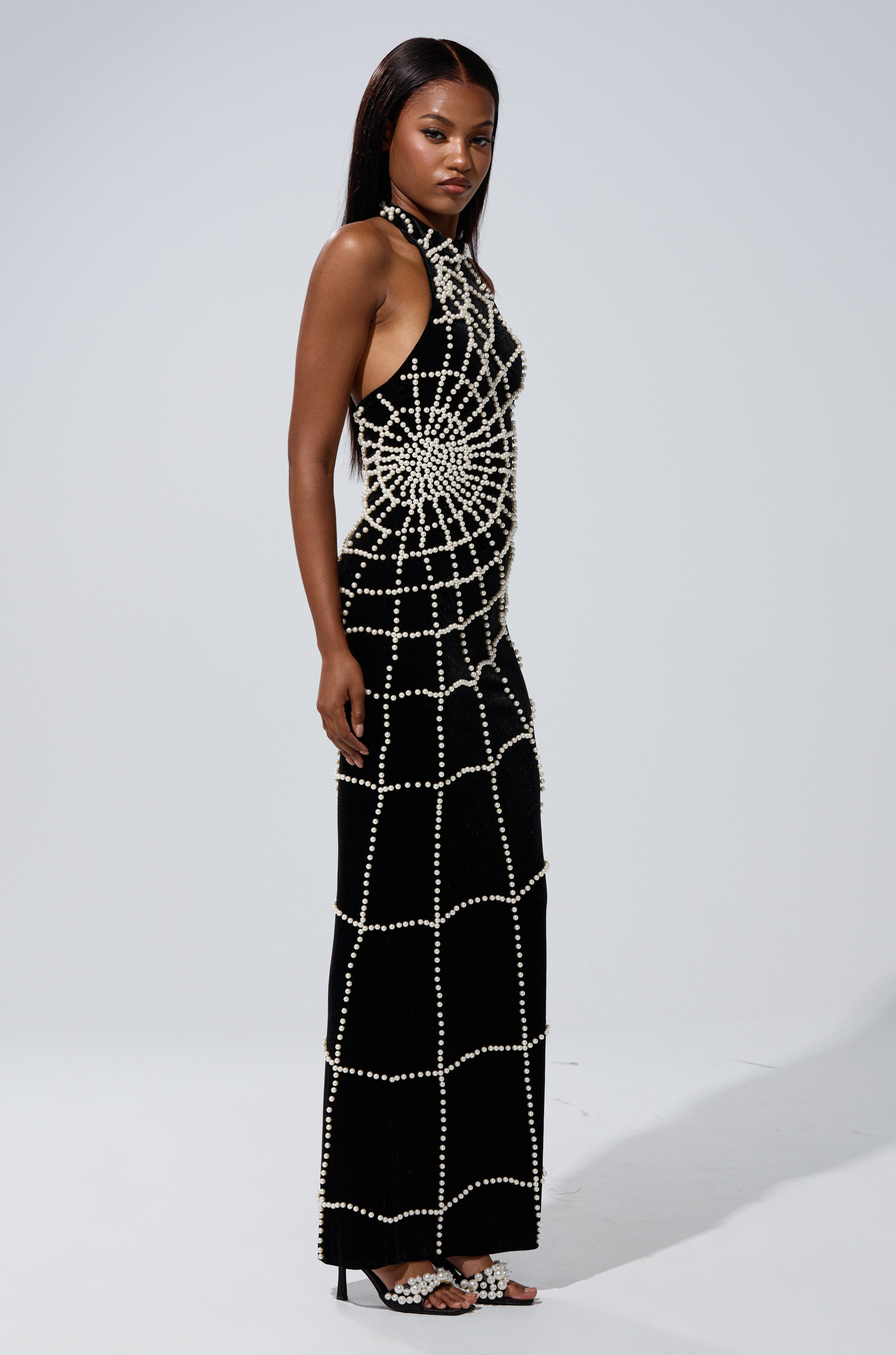 A woman exudes confidence in the MARYJANE MAXI DRESS WITH PEARLS, featuring a striking web of pearl embellishments, paired with matching pearl sandals. Her sleek dark hair completes the sophisticated ensemble.