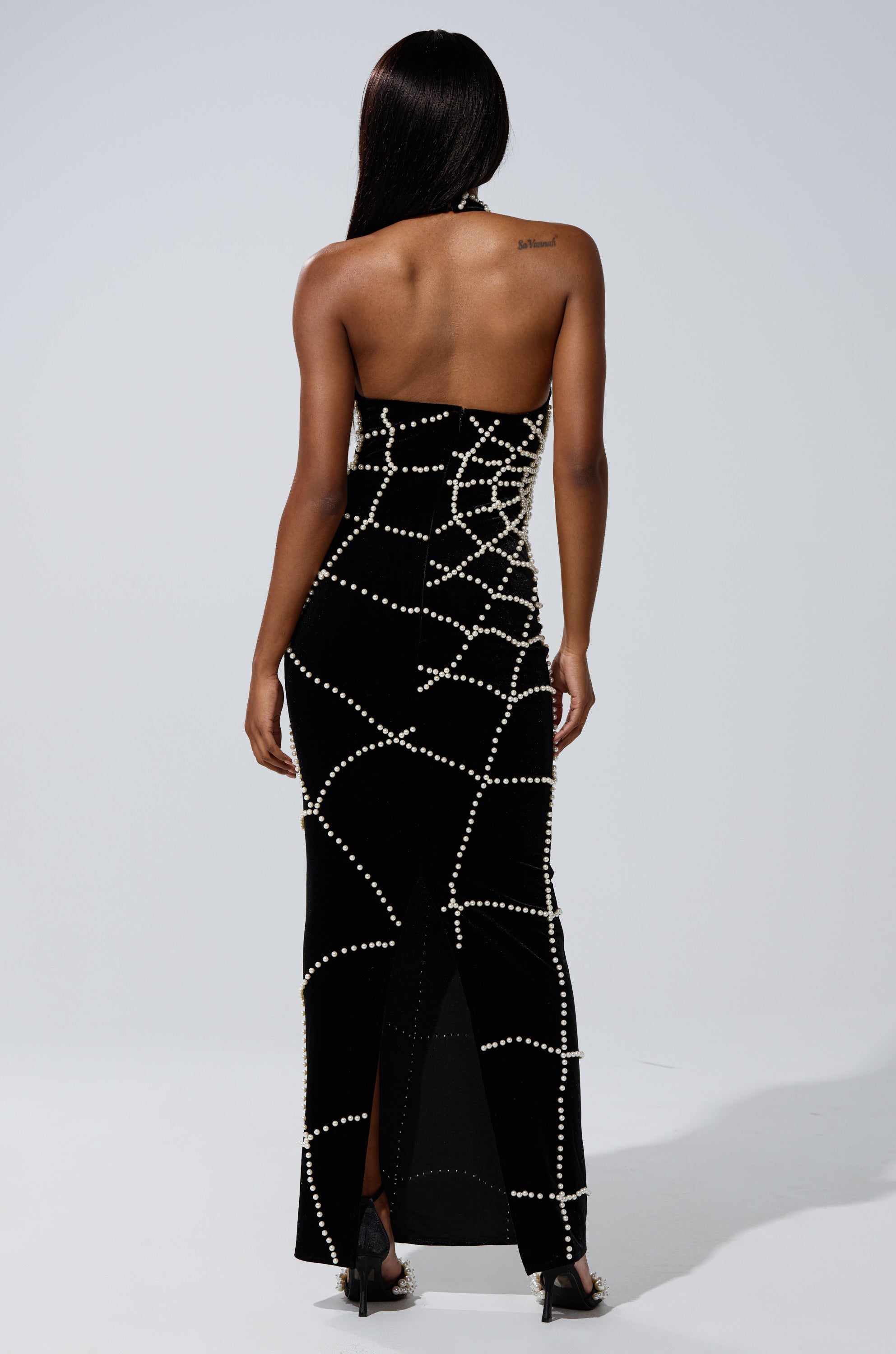 A woman faces away, wearing the MARYJANE MAXI DRESS WITH PEARLS—a strapless black velvet dress with a fitted silhouette and intersecting pearl embellishments—standing before a plain light background.