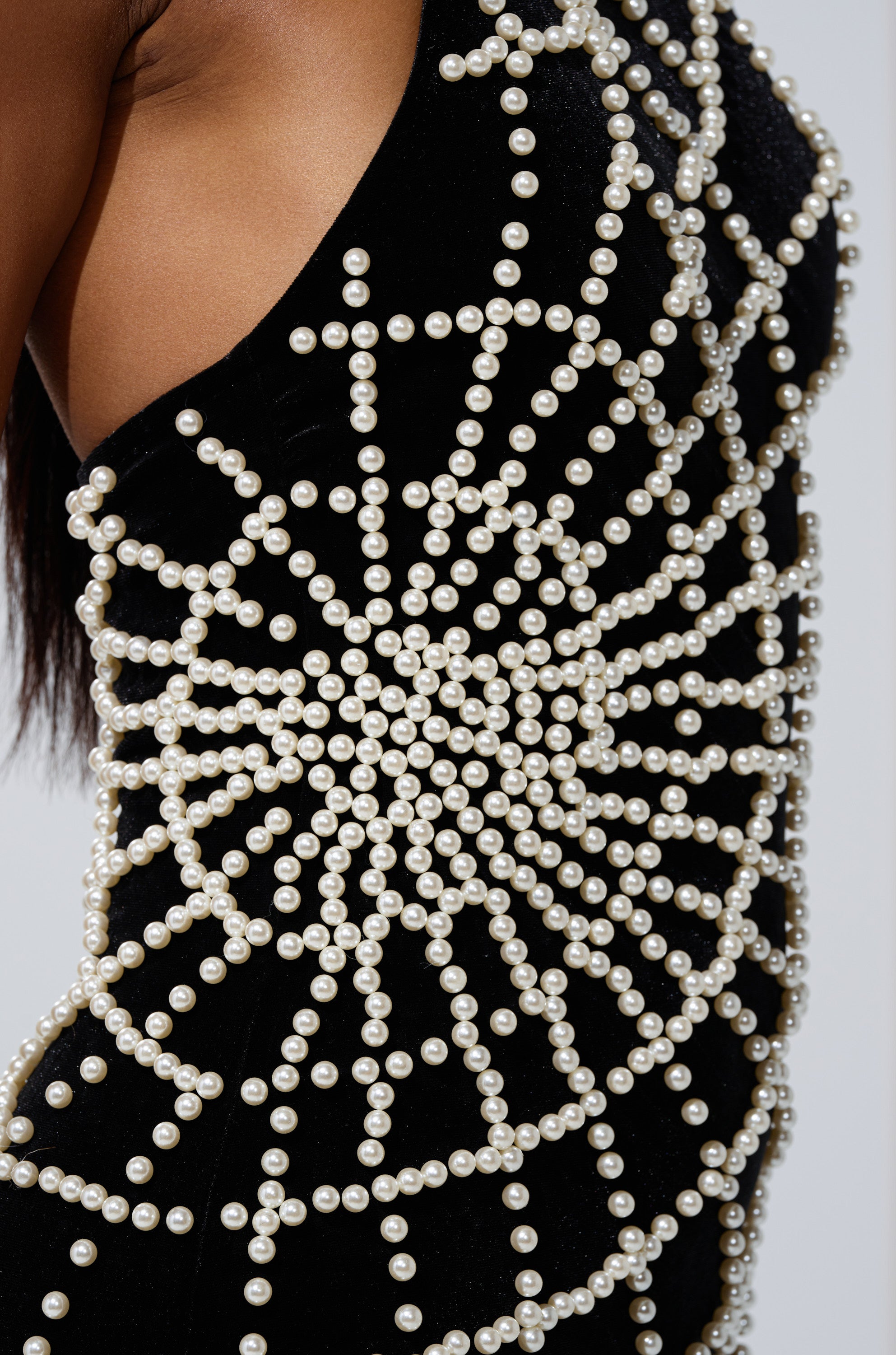 A close-up of the MARYJANE MAXI DRESS WITH PEARLS—a black velvet dress adorned with pearl accents in a radiant geometric web pattern, shown worn with part of the wearer’s arm and side visible.