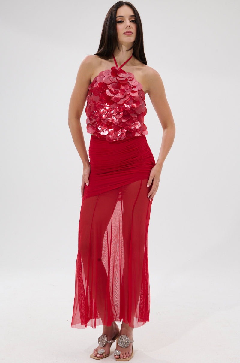A woman stands in a studio wearing the QUEEN OF EVERYTHING HALTER TOP, a sleeveless red statement piece with large sequin discs on the bodice. She pairs it with a sheer, flowy skirt, has long straight brown hair, and wears sandals.