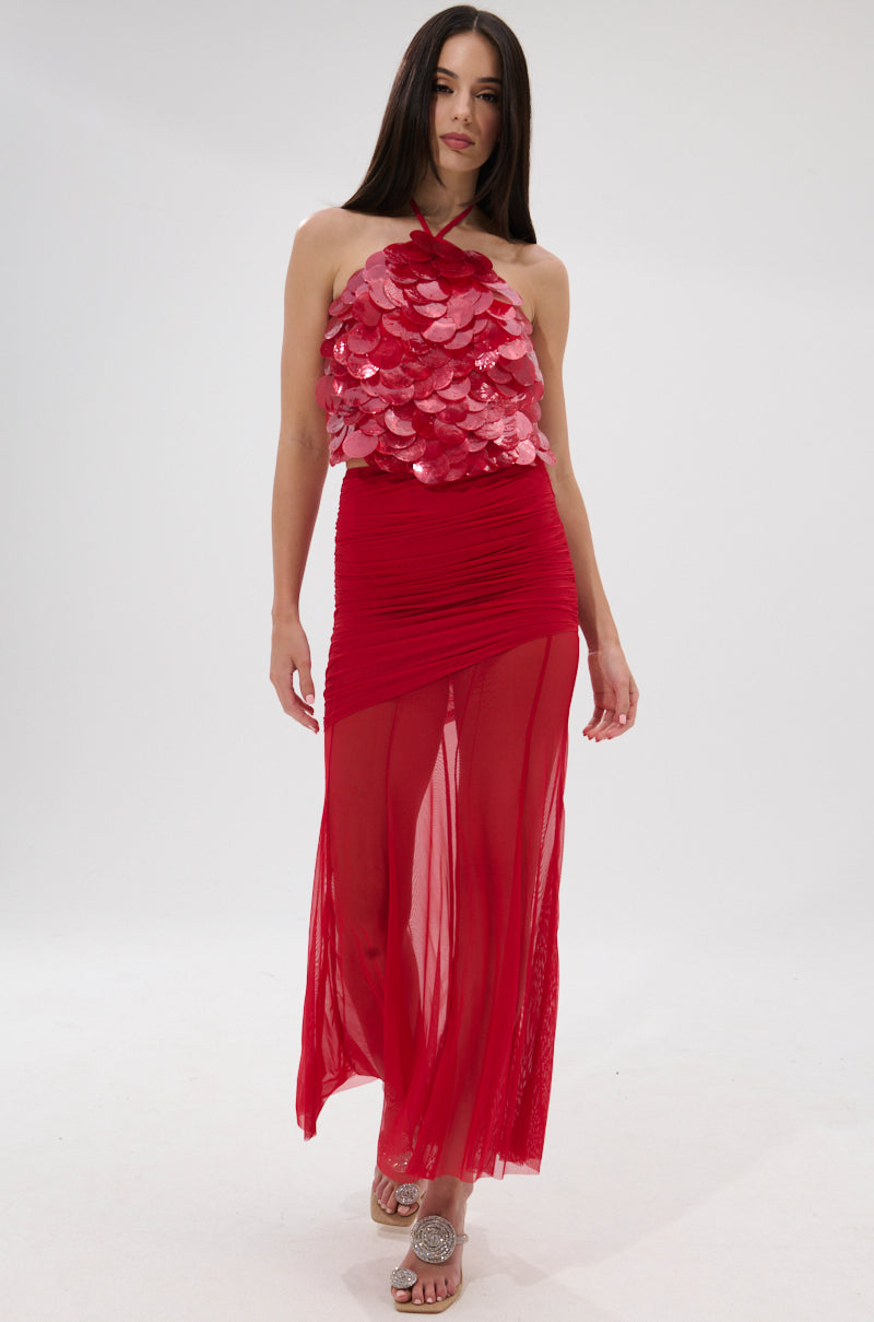 A woman stands against a plain white background wearing the QUEEN OF EVERYTHING HALTER TOP, a sleeveless red statement piece with shiny floral appliqués and a sheer, ruched skirt. She has long straight hair and is wearing sandals.