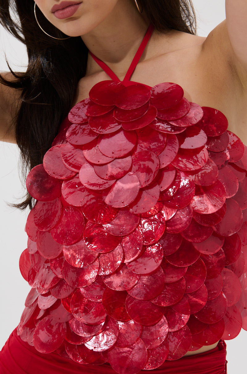 A woman in the QUEEN OF EVERYTHING HALTER TOP, featuring large shiny red sequins in overlapping layers, with long dark hair and hoop earrings.