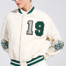 A person in a BETTER LUCK NEXT TIME EMBELLISHED BOMBER, an oversized white varsity jacket with green striped accents and the number "19" adorned on it. The sleeves feature pearl rhinestone details. They also wear sunglasses, a light pleated skirt, and have neatly styled blonde hair.