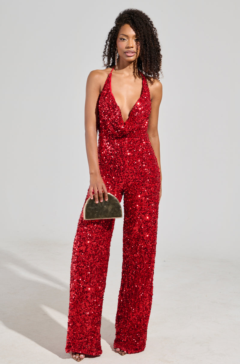 A person with curly hair showcases the Jodie Scoop Neck Sequin Jumpsuit in red, holding a small metallic clutch. They stand confidently against a plain background, casting a shadow on the floor.