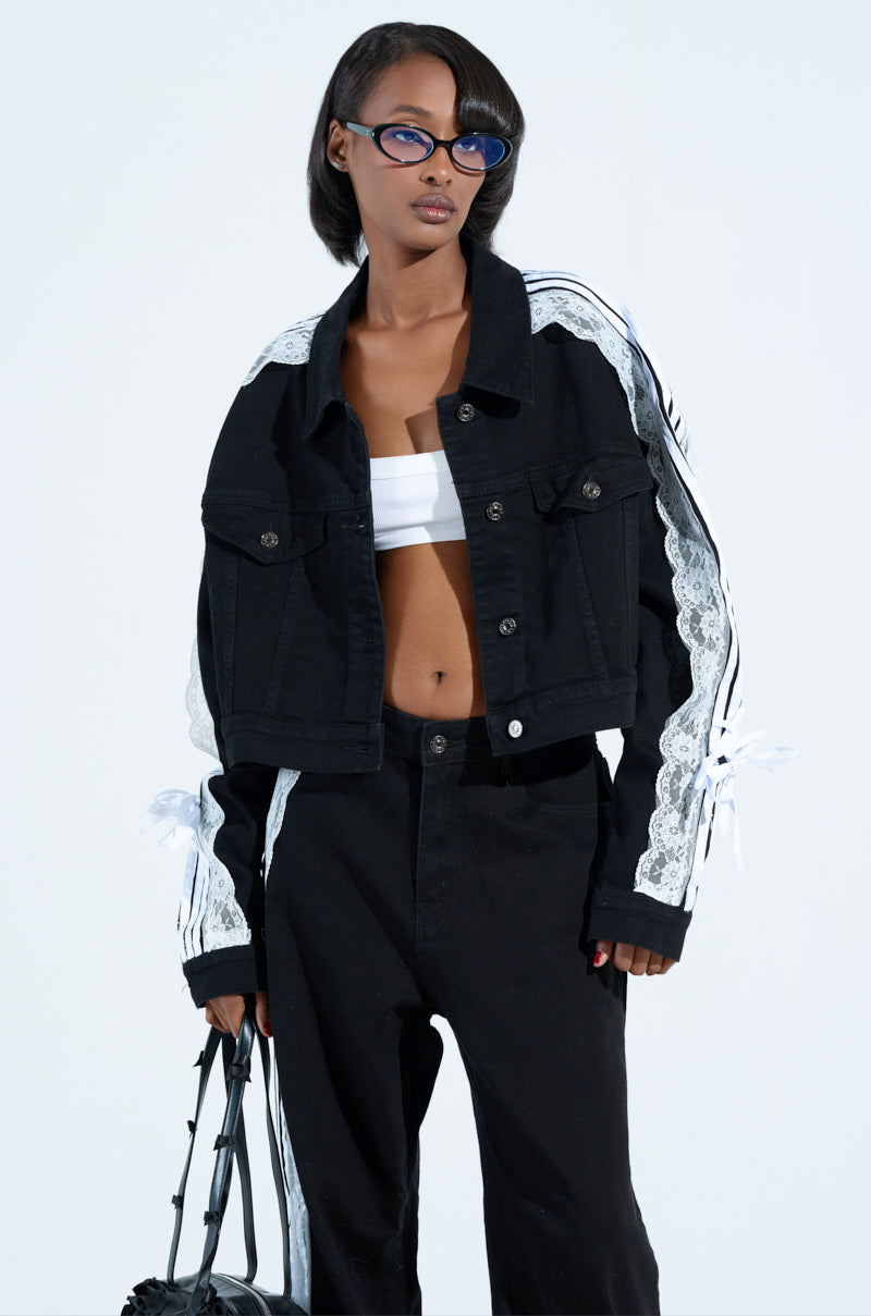 A person with bobbed black hair and blue glasses dons a Dylan Lace Detail Denim Jacket in dark wash with matching pants, a white bandeau top beneath. They hold a black bag. The ensemble has metallic button cuffs, all set against a plain white background.