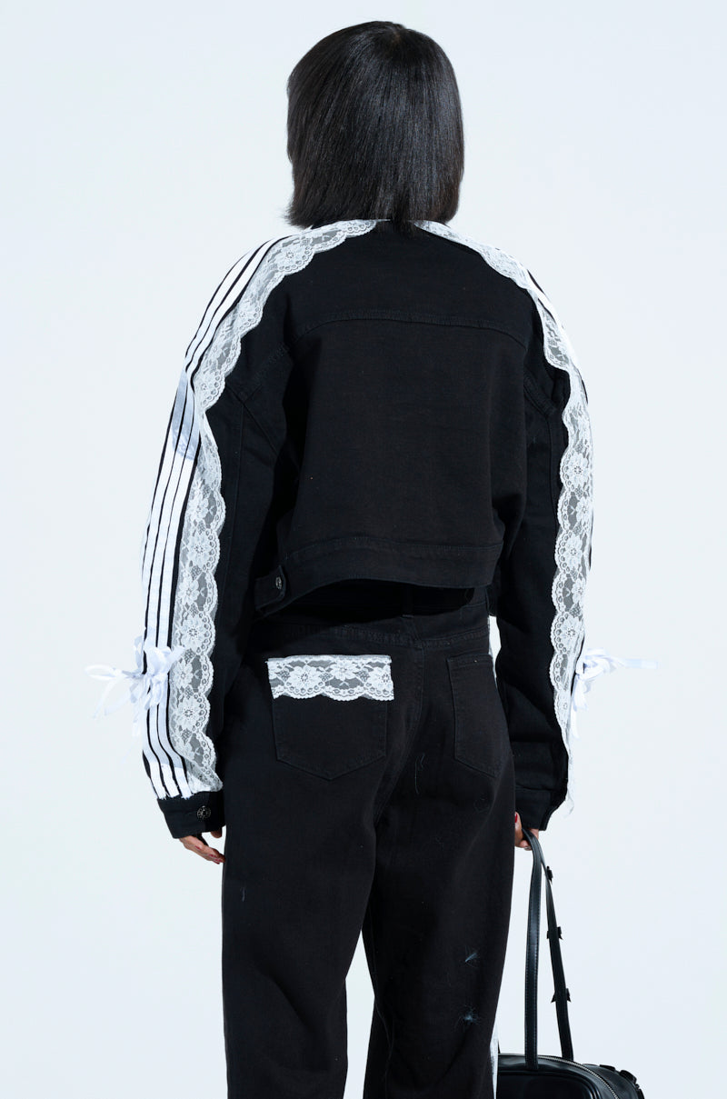 Someone with medium hair wears a Dylan Lace Detail Denim Jacket featuring metallic button cuffs and white lace stripes on the sleeves, paired with black pants having lace-trimmed back pockets, and carries a chic black bag.