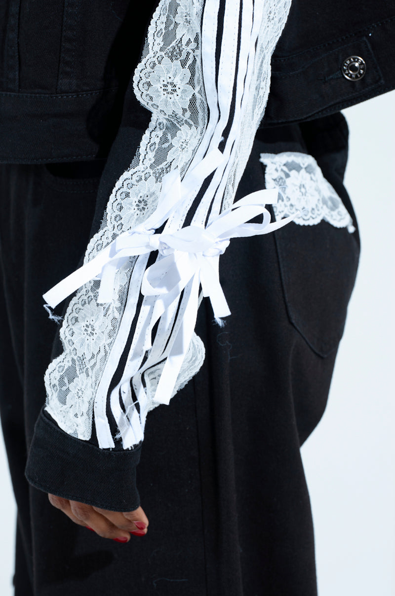 The individual wears a black Dylan Lace Detail Denim Jacket, highlighted by metallic button cuffs and white lace with ribbon bows. Red nails against black pants complete this effortlessly chic look, distinguished by eye-catching details.
