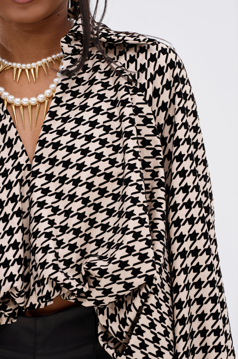 A close-up of a person wearing the I KNOW WHAT I WANT HOUNDSTOOTH BLOUSE TOP, layered gold and pearl necklaces, and black bottoms, with focus on the upper torso and clothing details.