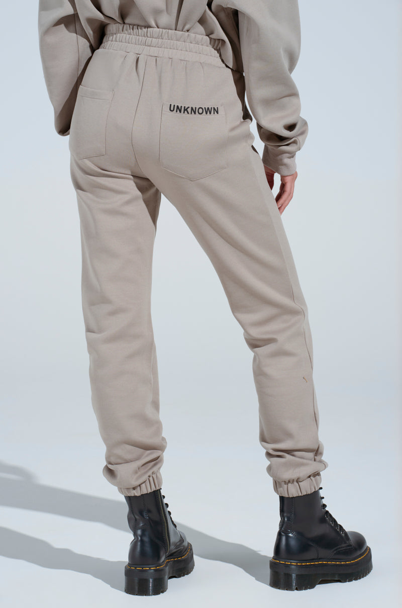 Person wearing light beige SOURCE UNKNOWN JOGGER pants, crafted from a soft cotton blend with "UNKNOWN" on the back pocket, paired with a matching sweatshirt and black lace-up boots, standing against a plain light background.
