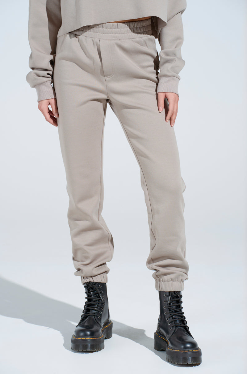 A person wears light beige SOURCE UNKNOWN JOGGER pants, a matching sweatshirt, and black lace-up combat boots while standing against a plain light background. The image is cropped at the shoulders.
