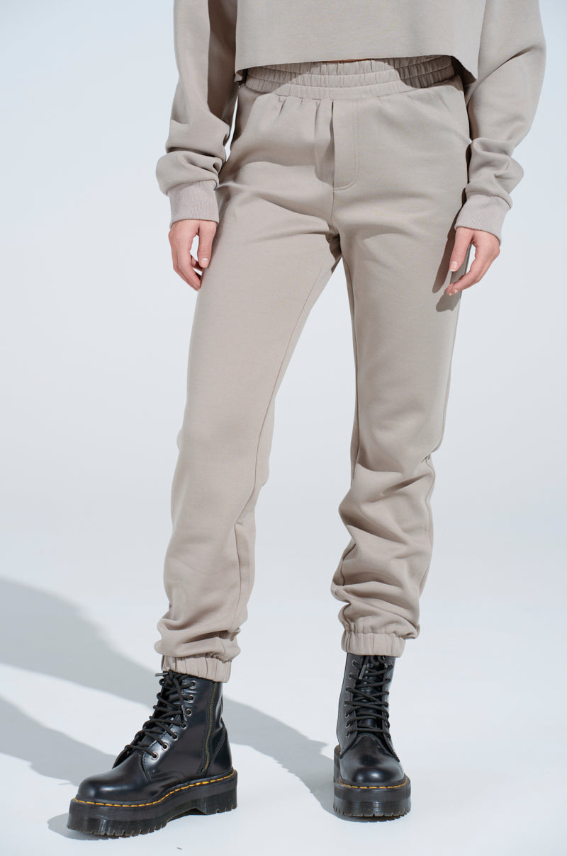 Person wearing beige SOURCE UNKNOWN JOGGER pants, a matching beige sweatshirt, and black lace-up combat boots, standing against a plain light background. The photo is cropped at the shoulders.