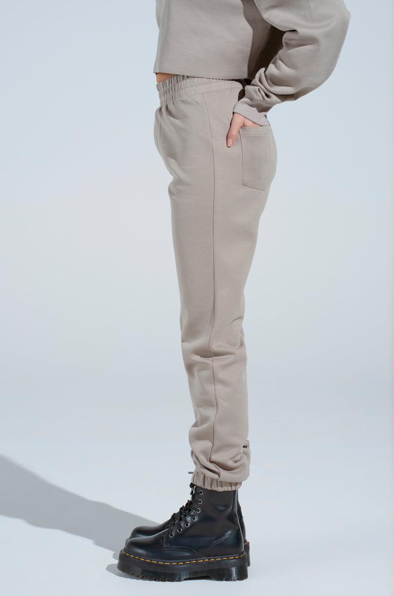 A person stands in profile wearing light beige SOURCE UNKNOWN JOGGER pants, a matching sweatshirt, and black chunky lace-up boots against a plain light background.