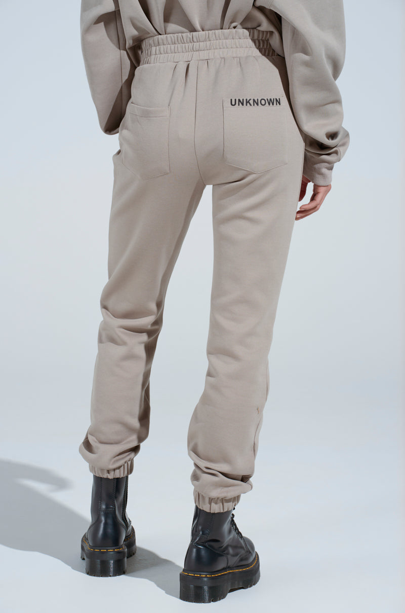 Someone stands against a light background wearing SOURCE UNKNOWN JOGGER high-rise pants with "UNKNOWN" on the back pocket, paired with black boots and a matching beige sweatshirt.