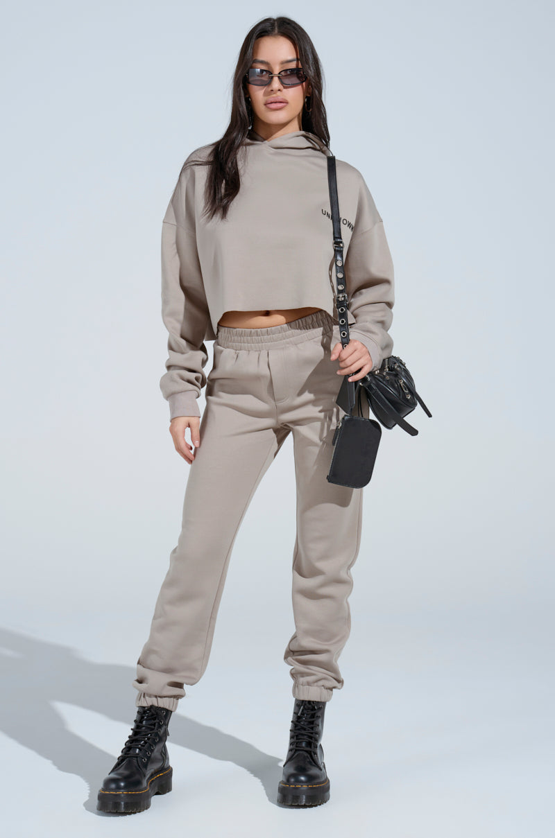 A woman in sunglasses wears a beige SOURCE UNKNOWN JOGGER set with a cropped hoodie and black combat boots, carrying a black crossbody bag. She stands confidently against a plain light background.