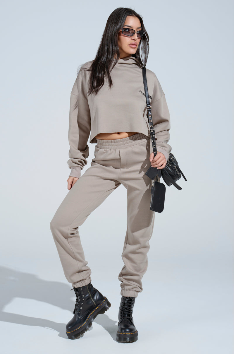 A woman in the SOURCE UNKNOWN JOGGER set—taupe cropped sweatshirt and high-rise jogger pants—stands confidently. She has long dark hair, sunglasses, black combat boots, and a small black crossbody bag against a plain light background.