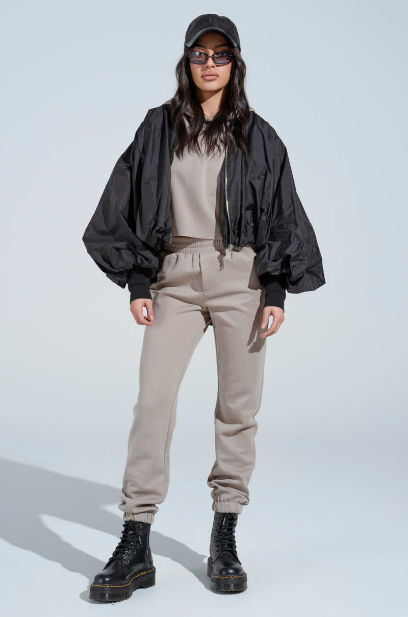A woman wearing a black cap, sunglasses, black bomber jacket, SOURCE UNKNOWN JOGGER beige cotton-blend jogger and sweatshirt set, and black lace-up boots stands facing forward against a plain light background.