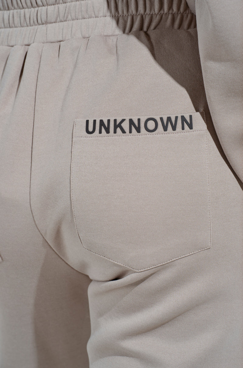 Close-up of beige SOURCE UNKNOWN JOGGER high-rise pants with an elastic waistband and a back pocket labeled "UNKNOWN" in black above the pocket.