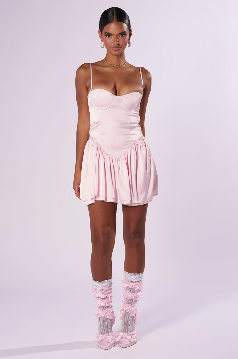 A woman stands against a plain background wearing the FELT CUTE SATIN MINI DRESS, a light pink spaghetti strap mini dress featuring a corset-inspired bodice and a flared skirt. She complements her outfit with matching pink ankle-high, ruffled socks and matching shoes. Her hair is pulled back, and she accessorizes with hoop earrings.